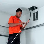 Technician in orange shirt working on wiring through ceiling in a white room, using tools to strip cables.