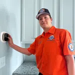 Technician in orange Estes Services uniform adjusting a thermostat set to 76 degrees in a kitchen.