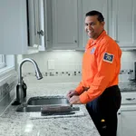 Smiling plumber in orange uniform standing in modern kitchen with granite countertops and sink tool on counter
