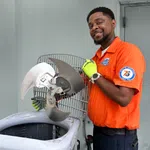 HVAC technician in orange uniform holding a dirty fan blade from an air conditioning unit during maintenance.