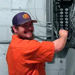 Smiling electrician working on a circuit breaker panel wearing an orange shirt and a branded cap indoors