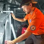 Technician in an orange shirt working on HVAC equipment with headlamp in an industrial setting.