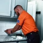 Electrician in orange shirt installing electrical outlet in modern kitchen with gray cabinets and granite countertop.