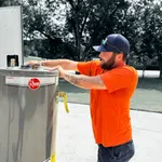 Technician in orange shirt installs or inspects Rheem water heater outdoors on sunny day with trees in background.