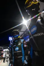 Race car driver in Konica Minolta suit and helmet stands in pit lane at night with bright lights and team members.