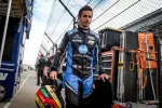 Race car driver in black and blue Konica Minolta racing suit holding a helmet walking in a pit lane.