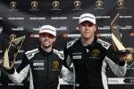 Two race car drivers in black Lamborghini racing suits holding trophies and posing on the winner's podium.