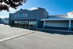 Gray storefront of At Home store with large windows and yellow bollards under a clear blue sky