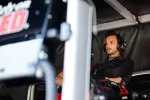 Focused race car driver wearing headset and black racing suit sitting with arms crossed in a pit area.