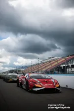Red race car leads a line of sports cars on a racetrack under heavy cloudy sky with empty grandstands.
