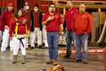 Group of workers in red shirts in a warehouse, one wearing a safety harness and others listening attentively.