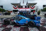 Blue and black Konica Minolta Acura race car with team celebrating victory on podium with confetti and banners.