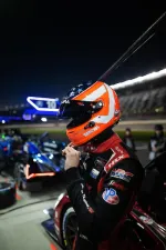 Race car driver in American flag helmet preparing at night pit stop with race cars and illuminated lanes in background