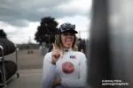 Woman in racing gear with cap and sunglasses sticking out tongue and holding up index finger outdoors.