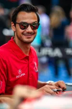 Smiling man wearing red Acura polo and sunglasses, signing autograph with blurred background crowd.