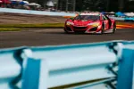 Red Acura race car speeding on a racetrack with blurred blue safety barrier in foreground.