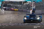 Blue Acura Konica Minolta race car leads competitors on a closed street circuit during a sunny race day.