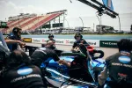 Race car pit crew members working on a blue IndyCar in the pit lane during a motorsport event with empty grandstands.