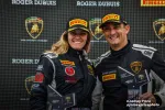 Two race car drivers in black Pirelli suits celebrating with second-place trophies at a Lamborghini event.