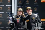 Two race car drivers in black racing suits with Pirelli caps hold trophies at a Lamborghini event backdrop.
