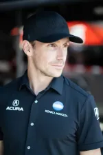 Man wearing black Acura and Konica Minolta branded shirt and black cap looking to the side with blurred background.
