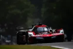 Red Acura race car with bright LED headlights racing on a dark track surrounded by trees.