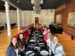Group of people smiling at a long dining table in a spacious room with wooden floors and exposed brick walls.