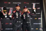 Two race car drivers in black racing suits celebrating with trophies on a podium at a racing event.