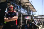 Race team crew member wearing headset and sunglasses standing with arms crossed at racing pit area.