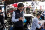 Man in Acura vest signing a helmet at a racing event booth with staff and visitors around.