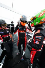 Race car driver adjusting helmet while team members in matching DEX Imaging suits prepare in garage pit area