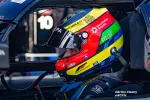 Race car driver wearing colorful helmet with sponsor logos inside a race car cockpit.