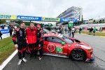 Race car drivers posing beside red number 93 sports car on the racetrack with Michelin banners overhead