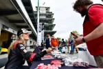 Race car driver signing autographs for fans at an outdoor motorsport event with pit building and crowd.