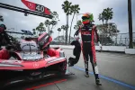 Race car driver in red and black suit walks beside Acura DPi race car number 40 in pit lane with palm trees.