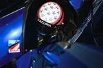 Race car driver wearing a star-patterned helmet with Acura branding inside a blue race car cockpit.