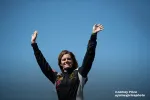 Smiling race car driver in black and white suit raises arms in victory against clear blue sky.