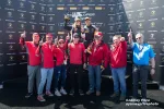 Winning racing team celebrating on podium holding trophy with team members showing number one gesture.