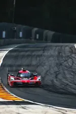 Red Acura race car speeding on a curved race track with tire marks and concrete barriers in the background