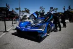 Blue Acura race car with open doors in pit lane surrounded by crew and equipment on a sunny day