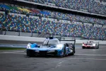 Blue and black Acura race car on a Daytona racetrack with colorful stadium seats and a red car in the background