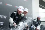 Race car drivers celebrating on the podium with champagne sprays wearing Hankook caps and racing suits.