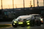 Race cars with bright yellow headlights compete on a track at dusk under a cloudy sky.