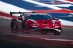 Red Lamborghini race car speeding on track with American flag blurred in background.