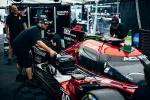Race car mechanics prepare a red and black Acura prototype race car in the garage area before a race event.