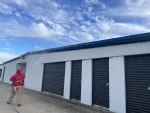 Person in red hoodie pressure washing white storage unit building with black doors under a blue cloudy sky.