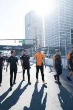 Group of race team members walking on city street near Chevrolet racing banners under bright sunlight.
