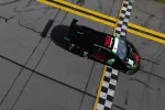 Black and red race car crossing the checkered finish line on asphalt track with yellow lane markings.