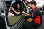 Two men interacting by a race car in a garage area, one seated inside the vehicle and the other crouching outside.