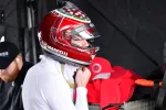 Race car driver wearing a red helmet and white suit preparing in the pit area with team members nearby.